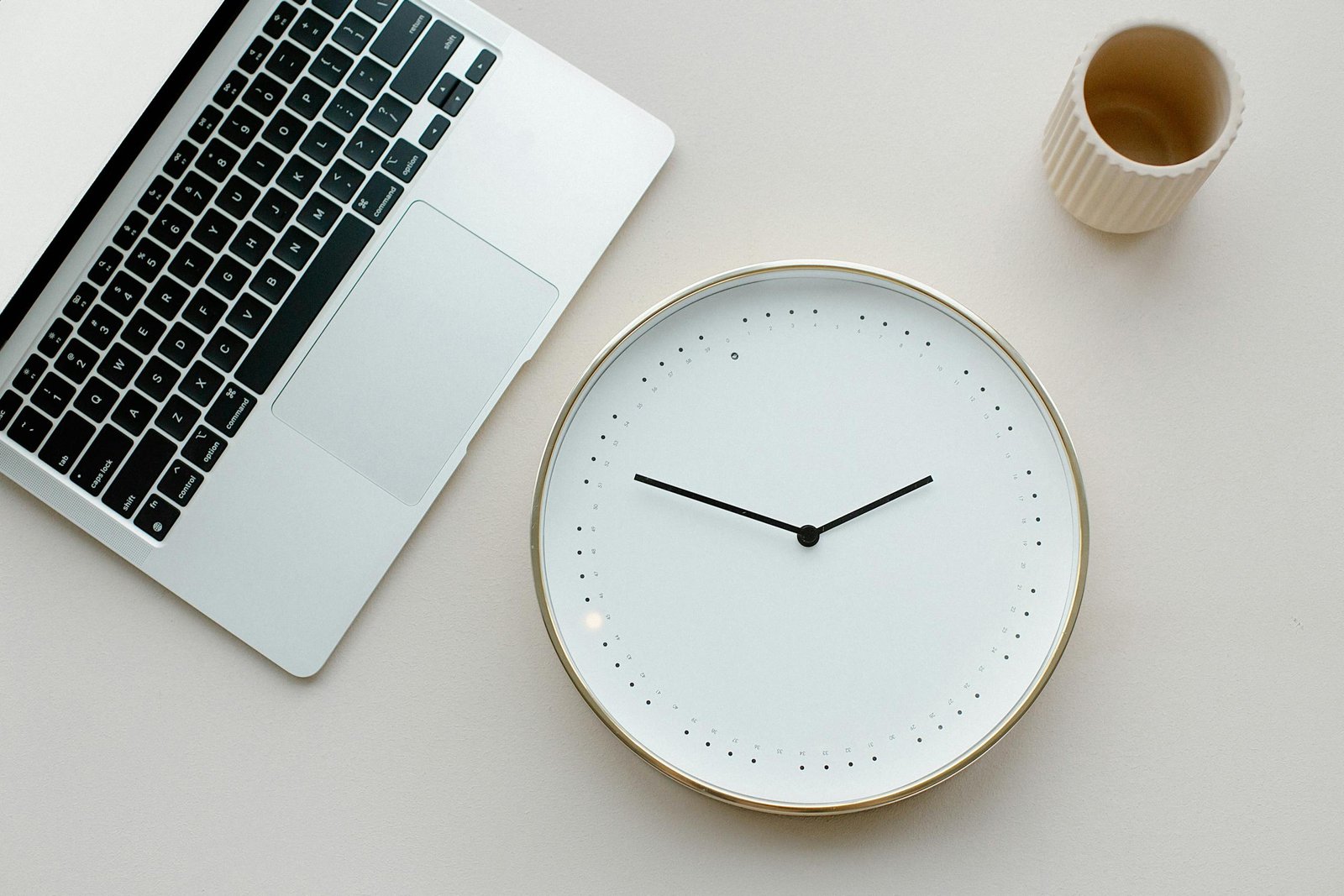 A clean minimalist workspace featuring a laptop, wall clock, and cup on a neutral background.