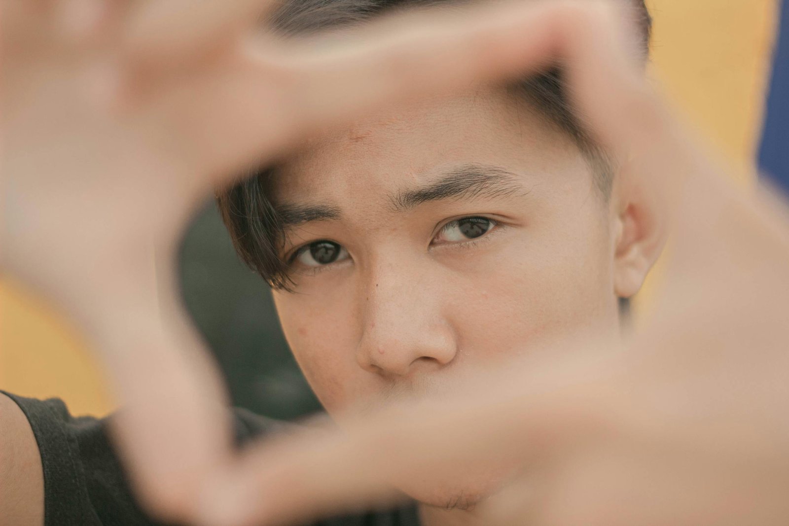 Close-up of a young man framing his face with hands outdoors, emphasizing eyes.